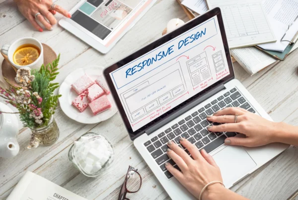 Person working on a laptop with a responsive web design layout displayed on the screen, surrounded by design materials including a tablet, a cup of tea, a book, and notes, illustrating the process of creating a responsive website.